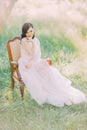 The full-lenght photo of the smiling woman in long white dress sitting on the old modern chair in the green spring field
