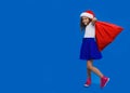 Full-lenght pretty little curly smiling girl wearing in santa hat and skirt carrying red bag, over on blue background. Royalty Free Stock Photo