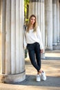 Full lenght of young woman in fashion clothing leaning against pillar outdoors. Royalty Free Stock Photo
