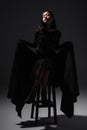 full length of asian model in black gothic dress and gloves sitting on high chair on dark grey,stock image Royalty Free Stock Photo