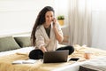 Full length of beautiful young lady using laptop and speaking on smartphone while sitting on bed at home, free space Royalty Free Stock Photo