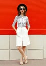 Full-length beautiful young woman model wearing white striped shirt, skirt, black round hat on city street over red wall Royalty Free Stock Photo