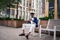 Full length black man sits on bench in urban environment and looks at phone, typing or browsing. Stylishly dressed guy