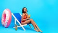 Full length of black woman drinking tropical cocktail in lounge chair, wearing swimsuit and laughing on blue background Royalty Free Stock Photo