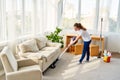 Full length body portrait of young woman in white shirt and jeans cleaning carpet with vacuum cleaner in living room, copy space.