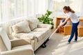 Full length body portrait of young woman in white shirt and jeans cleaning carpet with vacuum cleaner in living room, copy space.