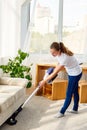 Full length body portrait of young woman in white shirt and jeans cleaning carpet with vacuum cleaner in living room, copy space. Royalty Free Stock Photo