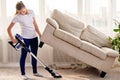 Full length body portrait of young woman in white shirt and jeans cleaning carpet with vacuum cleaner under sofa in living room. Royalty Free Stock Photo