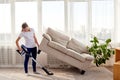 Full length body portrait of young woman in white shirt and jeans cleaning carpet with vacuum cleaner under sofa in living room. Royalty Free Stock Photo