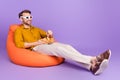 Full length body size photo of guy sitting in beanbag eating chips watching movie in 3d glasses vibrant violet