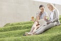 Full length of businesswomen with disposable coffee cup looking at laptop while sitting on grass steps