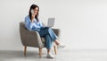 Full length of Caucasian lady using laptop and speaking on smartphone while sitting in armchair against white wall Royalty Free Stock Photo