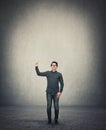 Full length contented businessman pointing forefinger up, standing in a room with grey wall background, empty copy space above