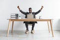 Full length of excited young black businessman sitting at desk with laptop, making YES gesture against white studio wall Royalty Free Stock Photo