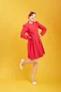 Full length of flirtatious woman with radiant smile wearing red dress with hand on hip isolated on yellow background