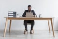 Full length of focused young black man working online with laptop computer, sitting at desk against white studio wall Royalty Free Stock Photo