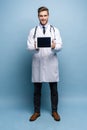 Full length of friendly doctor presenting empty digital tablet screen. Isolated on light blue. Royalty Free Stock Photo