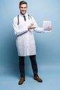 Full length of friendly doctor presenting empty digital tablet screen. Isolated on light blue. Royalty Free Stock Photo