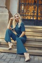 A full-length girl sits on the stairs in a long blue overalls. Blonde with gorgeous red make-up and dyno legs posing. Model on the