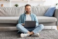 Full length of happy Caucasian man sitting cross legged on floor, using laptop pc, working online from home office Royalty Free Stock Photo