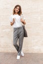 Full length image of beautiful modern woman standing against beige wall outdoor with silver laptop, and takeaway coffee in hands