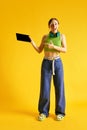 Full-length image of beautiful young woman in stylish clothes pointing on tablet screen against yellow studio background Royalty Free Stock Photo