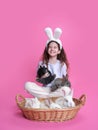 Full length view of a little girl with bunny ears, showing straw basket with little rabbits, on a pink background. Royalty Free Stock Photo