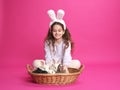 Full length image of a little girl with white bunny ears, showing straw basket with little rabbits, pink background. Royalty Free Stock Photo