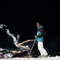 Male skier standing on snowy hill at night.