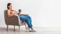 Full length of millennial woman drinking fresh coffee, having break in comfy armchair against white wall, panorama Royalty Free Stock Photo