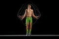 Full-length of muscular, athletic young man in motion, training, jumping rope against black background with stroboscope