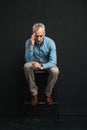 Full length photo of caucasian male pensioner 60s with short grey hair sitting on chair and looking down with brooding or uptight