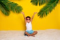 Full length photo of charming carefree small boy wear white shirt dark spectacles sitting sand rising hands arms