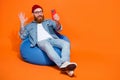 Full length photo of excited funny man wear jeans shirt sitting bean bag talking device empty space isolated orange