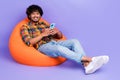 Full length photo of optimistic man dressed shirt sit in orange pouf hold smartphone read post on purple color