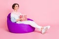 Full length photo portrait of woman with big popcorn bag sitting in violet beanbag chair isolated on pastel pink colored