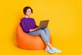 Full length photo portrait of woman working on laptop sitting in orange beanbag chair isolated on vivid yellow colored Royalty Free Stock Photo