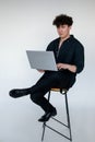 Full length photo of tired young curly guy in total black look sitting cross legged on chair, typing and working on Royalty Free Stock Photo