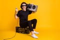 Full length photo of young man hold mic open mouth shoulder boombox sit wear black t-shirt pants white sneakers sunglass