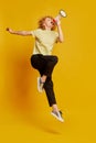 Full-length portrait of active young red-haired girl jumping with megaphone isolated on yellow studio background. Human Royalty Free Stock Photo