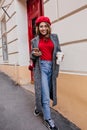 Full-length portrait of amazing woman in jeans and long coat comes out from coffee house with latte. Glad french girl in Royalty Free Stock Photo