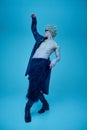 Full length portrait of artistic young man with naked torso in long black leather jacket dancing against blue studio Royalty Free Stock Photo