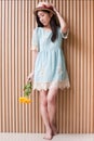 Full length portrait of asian girl touching hat and holding bouquet of yelliow flowers against wood wall