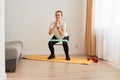 Full length portrait of athletic woman wearing white t shirt and black leggins, doing squats with resistance band on knees, Royalty Free Stock Photo