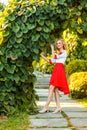 Full length portrait attractive happy blonde woman in stylish red white dress posing near floral arch in garden. standing and Royalty Free Stock Photo