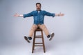Full length portrait of an attractive young man in jeans shirt sitting on the chair over grey background. Royalty Free Stock Photo