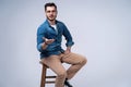 Full length portrait of an attractive young man in jeans shirt sitting on the chair over grey background. Royalty Free Stock Photo