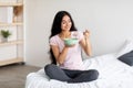 Full length portrait of beautiful millennial Indian lady having cereal with milk while sitting on bed at home Royalty Free Stock Photo