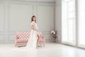 Beautiful pregnant young woman in white dress peignoir standing near pink sofa, holding with love her belly Royalty Free Stock Photo