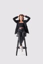 Full length portrait of beautiful young girl sitting on tall chair in black jeans and jacket over gray background Royalty Free Stock Photo